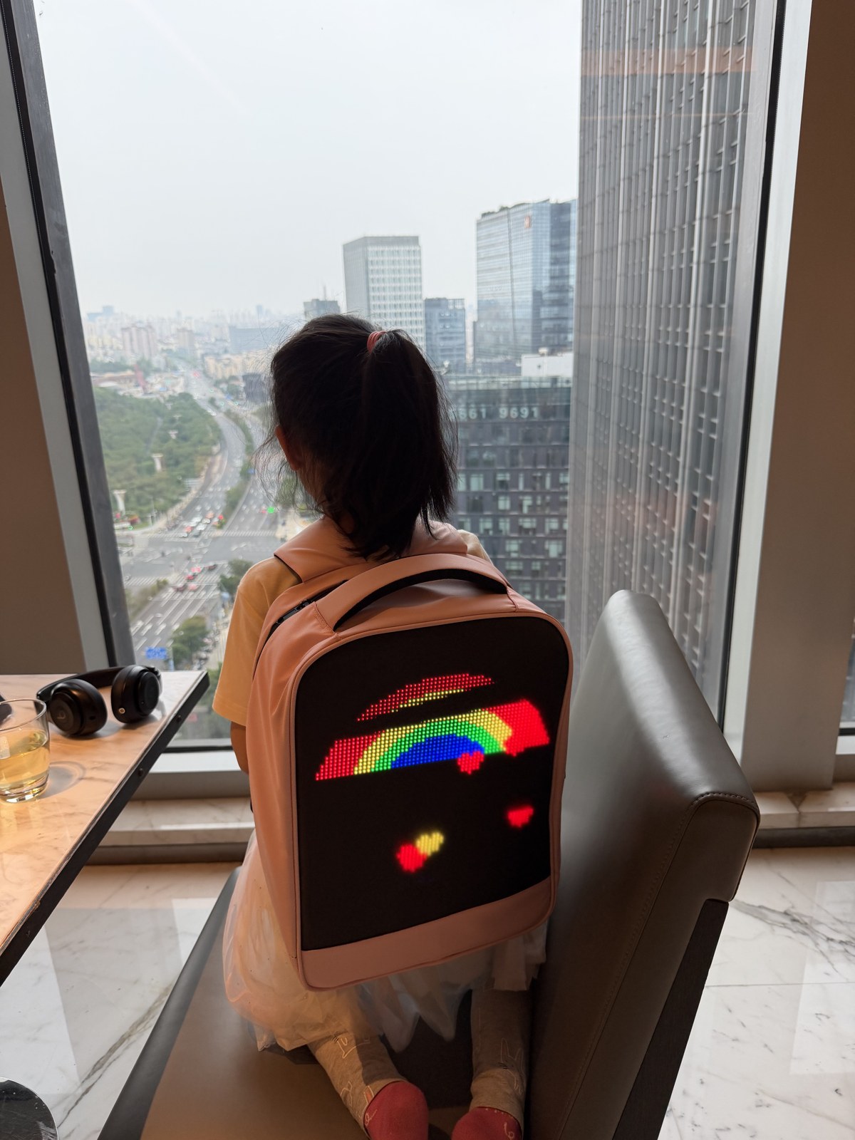 Child seen from behind, wearing a backpack whose LED panel shows a rainbow with floating hearts